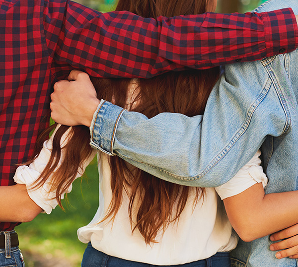 A rear shot of a female teenager with long hair who is walking in a park, is flanked either side by her parents/guardians who each have an arm around her. She has her arms around each of them too.