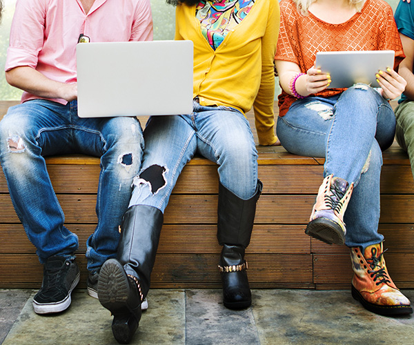 Three young people are sitting in a row on a wooden bench. The people on either side of the person in the middle both have open laptops.